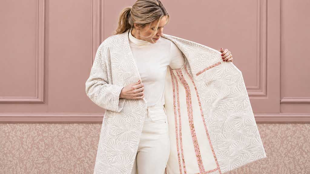 A woman wearing a cream sweater and pants holds a matching coat open to showcase the garment's clean interior construction and decorative seam finishing.