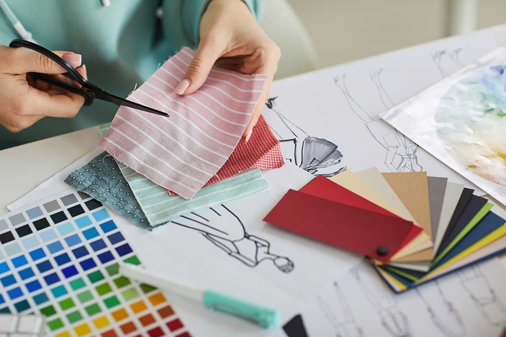 A person sitting at a desk cutting a fabric swatch. Understanding garment sourcing and fashion manufacturing is key when learning how to start a fashion brand. Building a fashion brand tech pack requires business partners across the supply chain. The owner of a fashion startup needs to understand industry terminology like fashion brand MOQ minimum order quantity and timelines as well as have the creativity to design garments. Studying how to find a fashion manufacturer will help ensure a good fit for your needs.