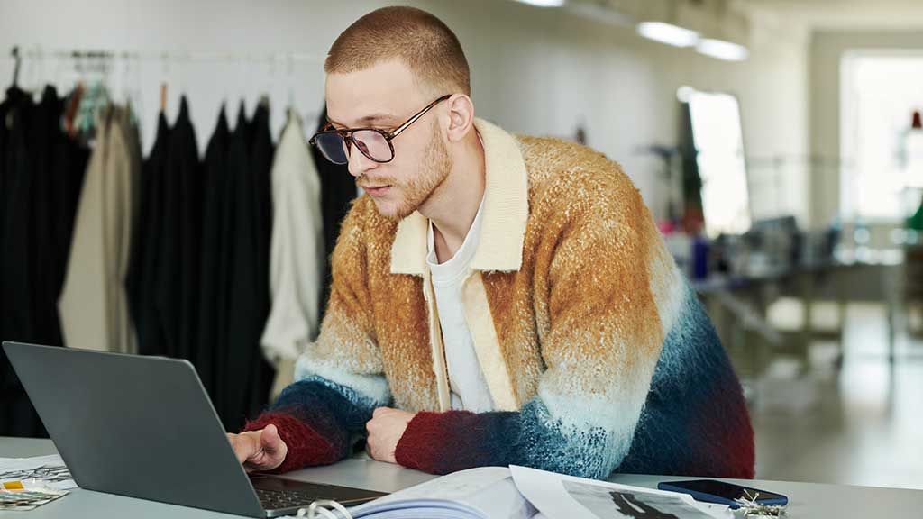 A man leans on a desk while working on a laptop in a fashion studio. He is searching how to write a sourcing inquiry. Clear and concise supplier communication shows apparel manufacturing partners you&rsquo;re prepared for a solid business relationship. Fashion suppliers work with brands that understand their MOQs and manufacturing capabilities, knowing how to contact a clothing manufacturer. Every brand&rsquo;s sourcing strategy should begin with understanding the details of the garment they want to produce and matching that with apparel manufacturers that meet those needs.