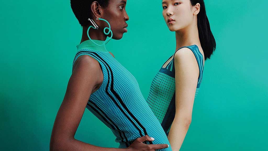 Two models pose against a teal background wearing form-fitting, sleeveless knit dresses in matching green tones. Computerized knitting machines are ubiquitous knit technology used to create the dresses.&nbsp; The knit fabrics have stripe and mesh patterns. Both models are in editorial poses, highlighting the garments&rsquo; innovative knit construction.