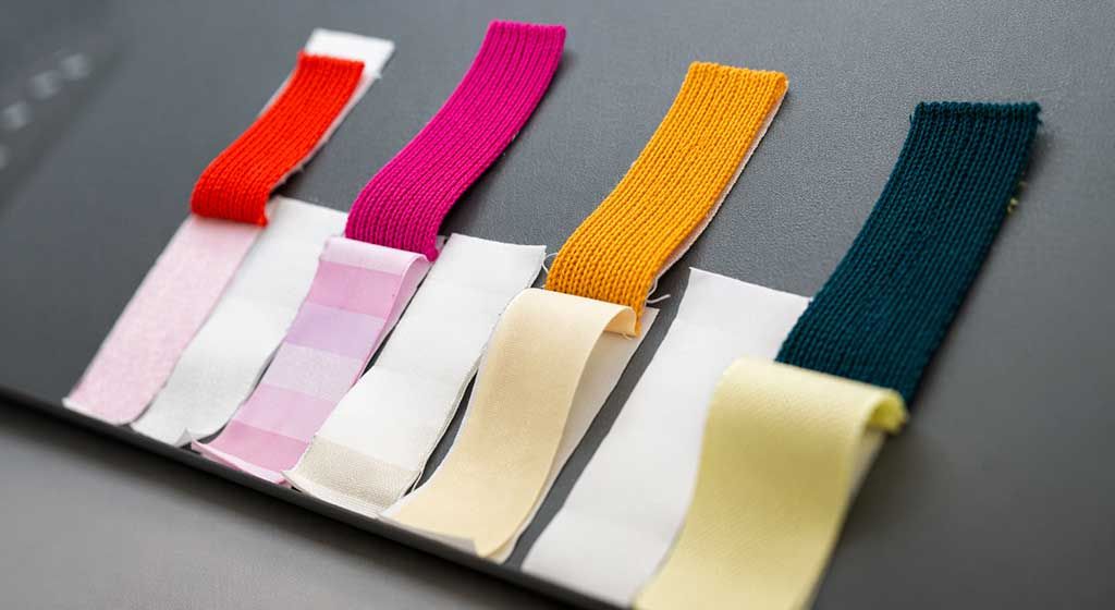 Fabrics in different colorways, including red, fuschia, yellow, and dark green being tested for colorfastness. Colorfastness can be measured by in-house testing or lab testing as part of fabric performance testing. The white strip of fabric to the right side of each color measures colorfastness, while the fabric immediately below tests for rating.