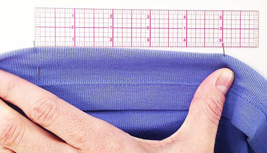 A 10 cm section of periwinkle jersey style knit fabric undergoing in-house fabric testing. The original fabric length is marked with pins for stretch testing. At 25% stretch, the far pin aligns with the 12.5-cm ruler mark. This test is a common measure of fabric performance and can be done with lab testing.