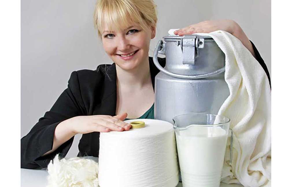 Blonde woman smiling with her hand on a metal milk can, In front, a glass jug of milk and a large spool of the sustainable fabric milk silk plus raw milk silk. 
