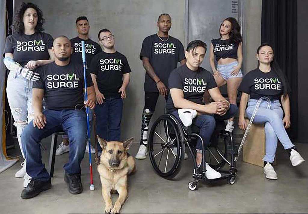 A group of racially diverse people with various physical disabilities wearing adaptive fashion. All wear Social Surge logo t-shirts and jeans as they sit/stand with their mobility aides and a seeing eye dog.