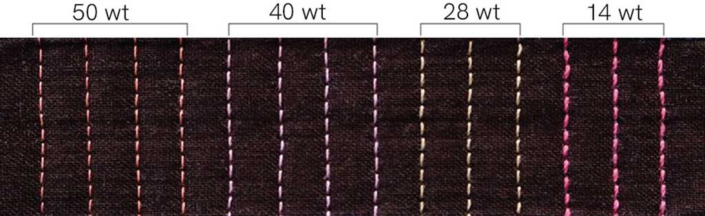 50, 40, 28, and 14 weight thread stitched vertically on a dark fabric. The 50 weight thread is peach and has the smallest diameter. The 40 weight thread is lavender. The 28 weight is yellow , 14 weight is hot pink with the largest diameter. There are four rows of stitching in each thread weight.